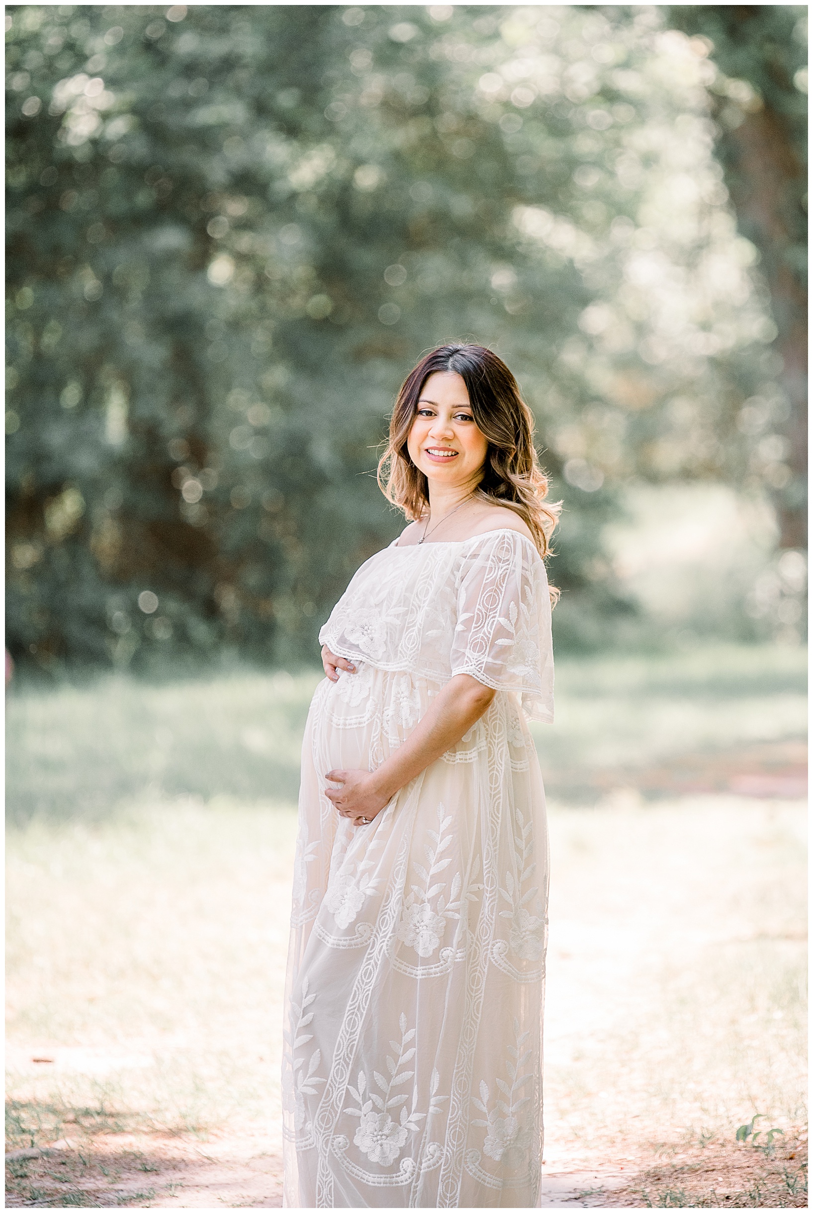 Woman in white embroidered maternity gown standing against backdrop of trees