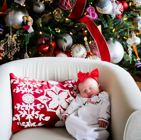 Baby in white onesie and red bow asleep in an armchair by the Christmas tree