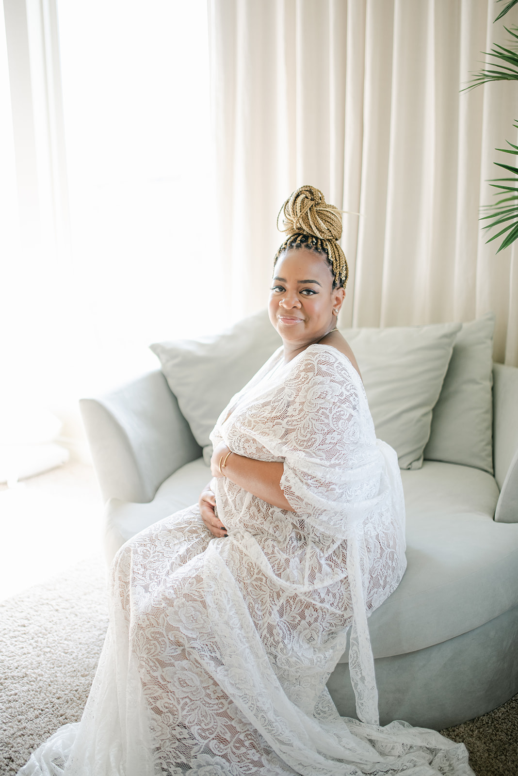 Black woman in white lace gown seated by window during maternity session in Texas