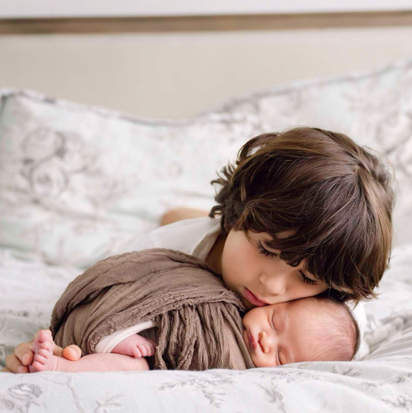 Little boy lying on bed and leaning his cheek against his new baby brother's head