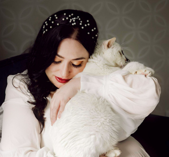 Dark-haired woman in white dress and pearl headband hugs her cat close, illustrating tips for including pets in your at home session