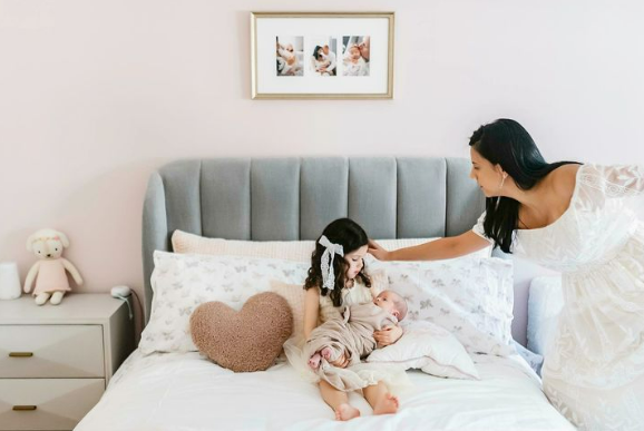 Your Houston newborn photography session at home, mother reaching out towards her daughter as the little girl sits on a bed holding her new baby sibling