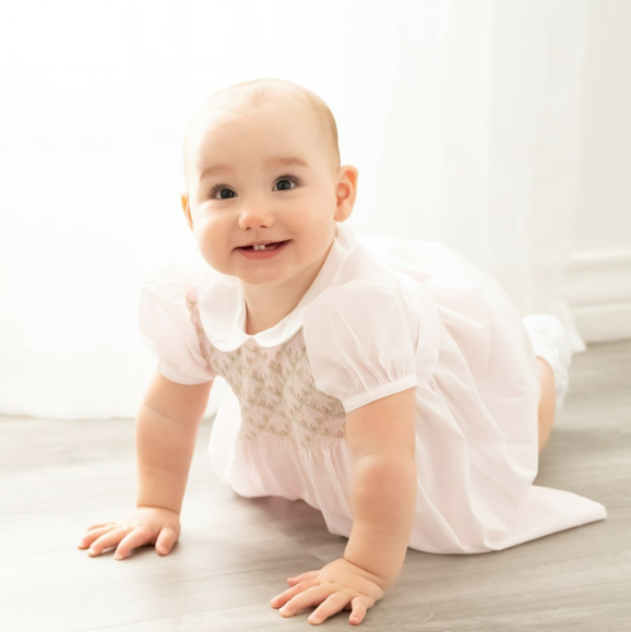 Smiling baby girl in white dress crawling across the floor of Houston TX studio