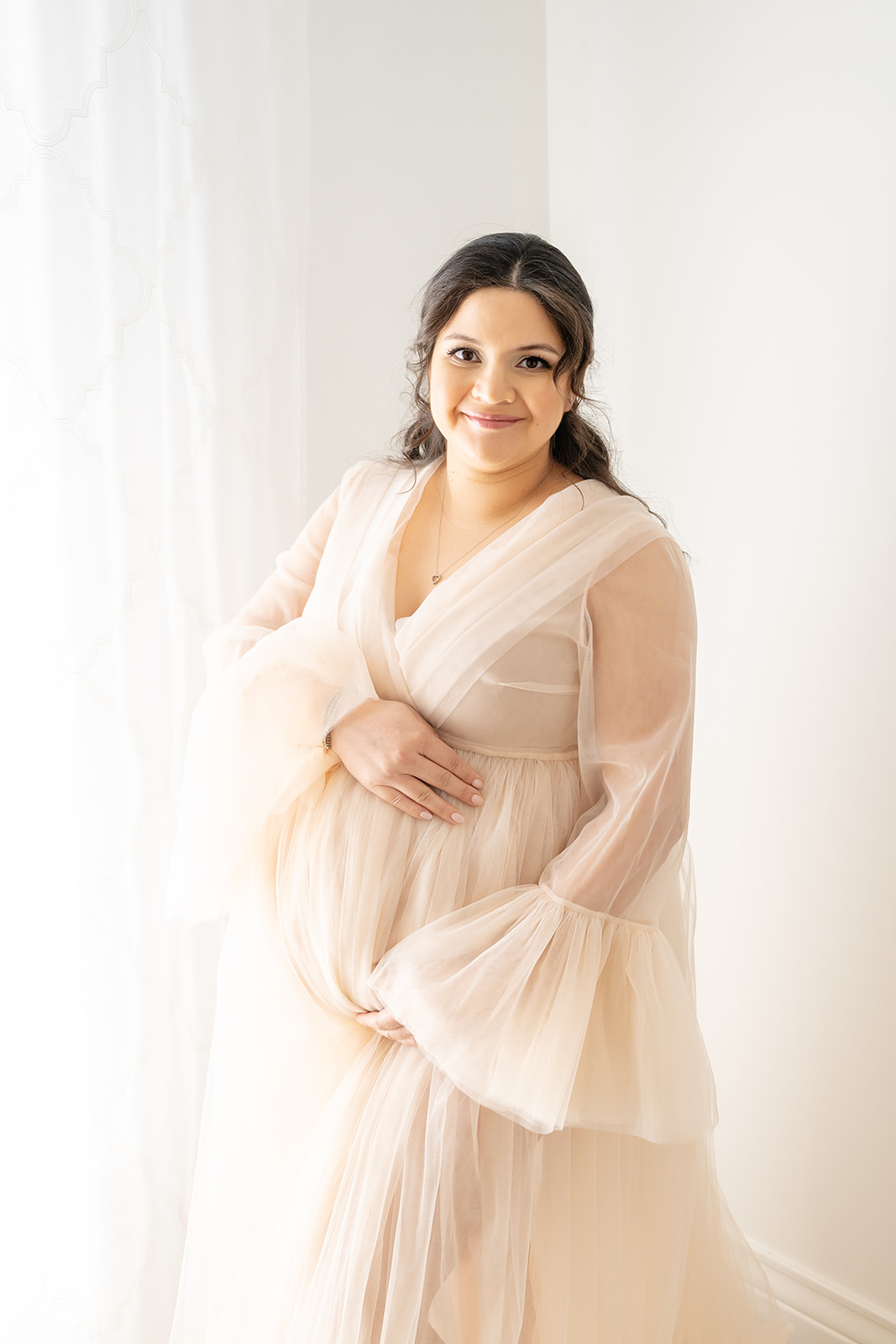 Smiling woman in creamy tulle maternity gown standing next to a window against a white backdrop