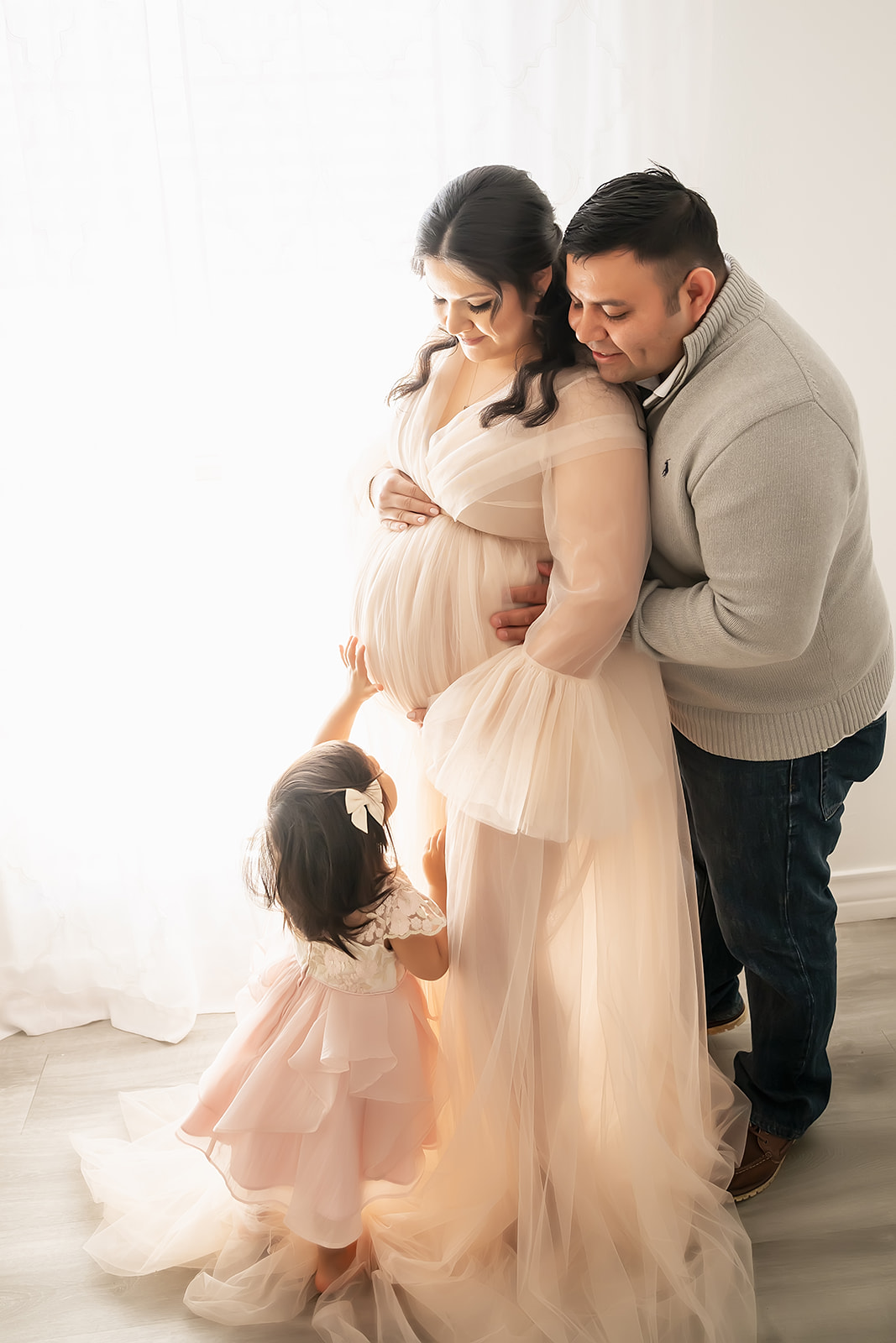 Toddler girl reaches up to touch mom's tummy as dad peeks over mom's shoulder during a studio maternity session with siblings in Houston Texas