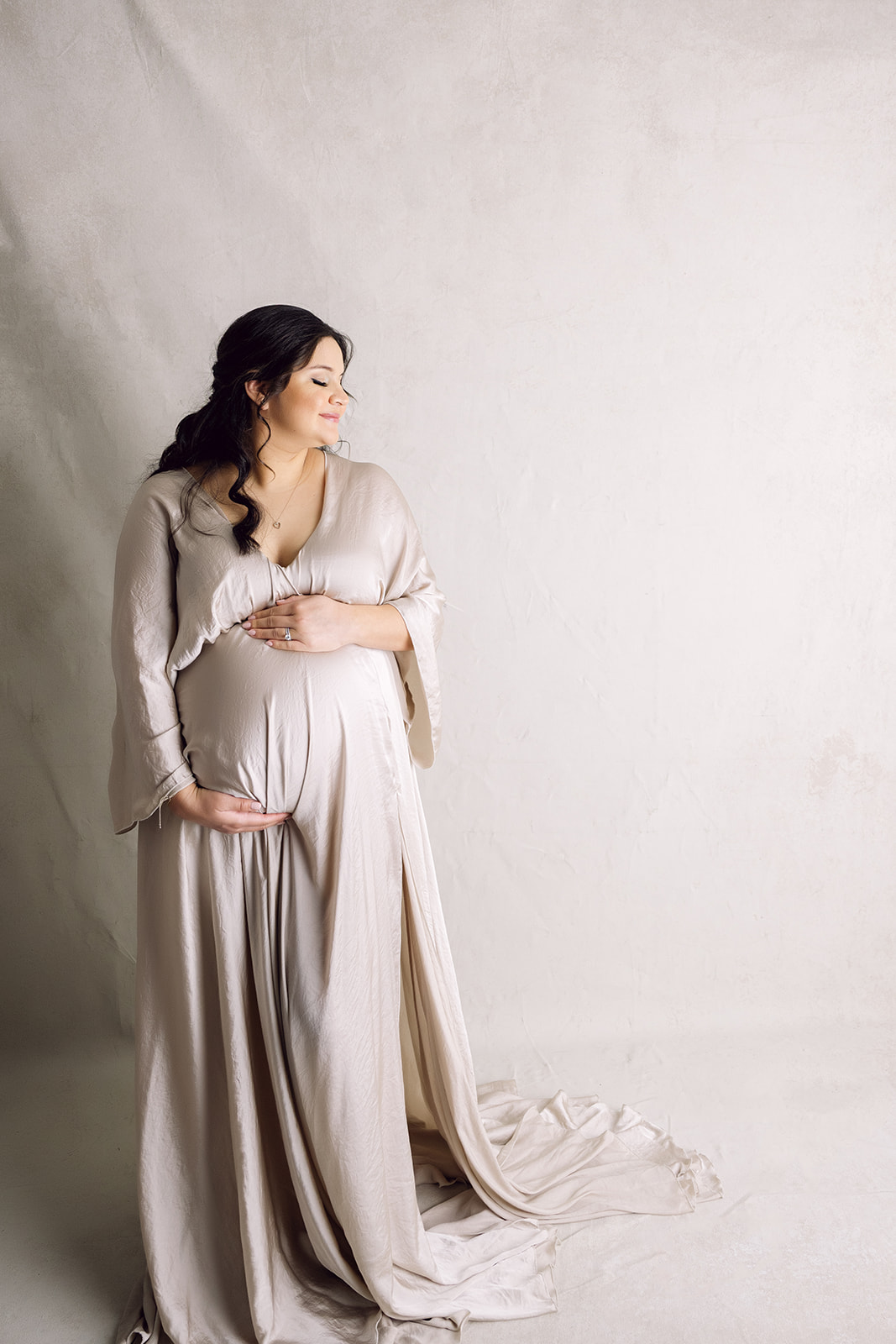 Woman in flowing satin maternity dress against an ivory background during her studio maternity session