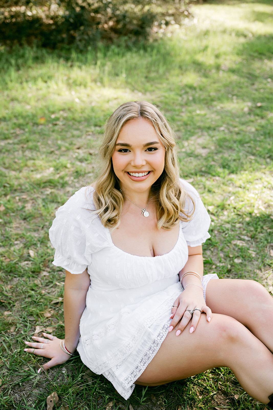 Young woman in white summer dress seated on the grass and smiling up at the camera during her girl's high school senior session