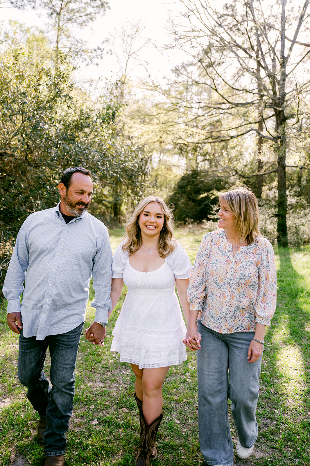 High school senior girl in white summer dress and boots walking hand in hand with her parents through a sunny park near Houston Texas