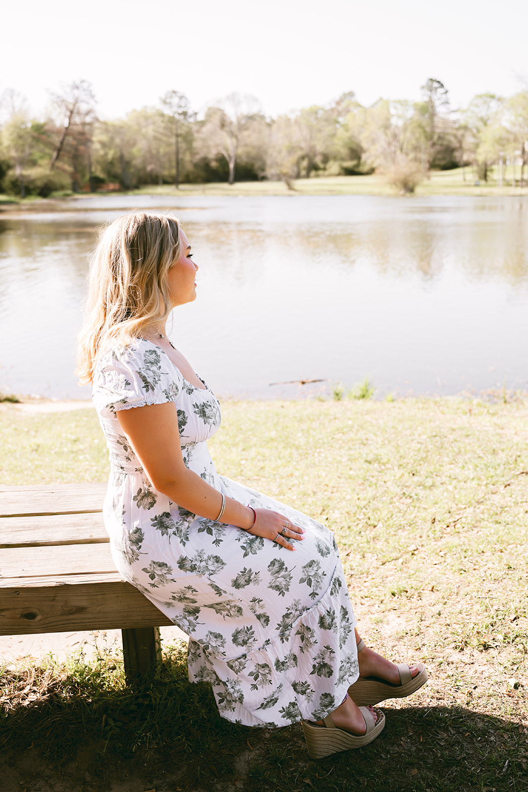 High school senior girl in white floral print dress seated on a wooden bench and looking out over a lake in Houston Texas