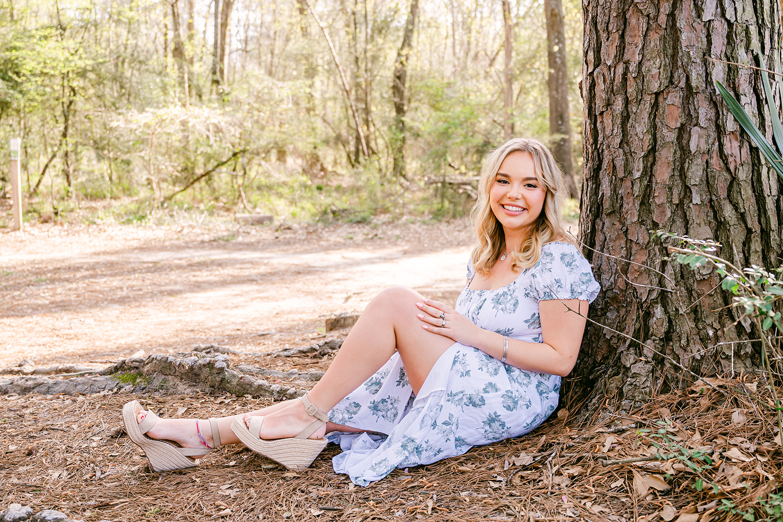 Young woman in blue and white floral print dress and wedge sandals seated on the ground leaning against a tree during her senior portraits in Houston