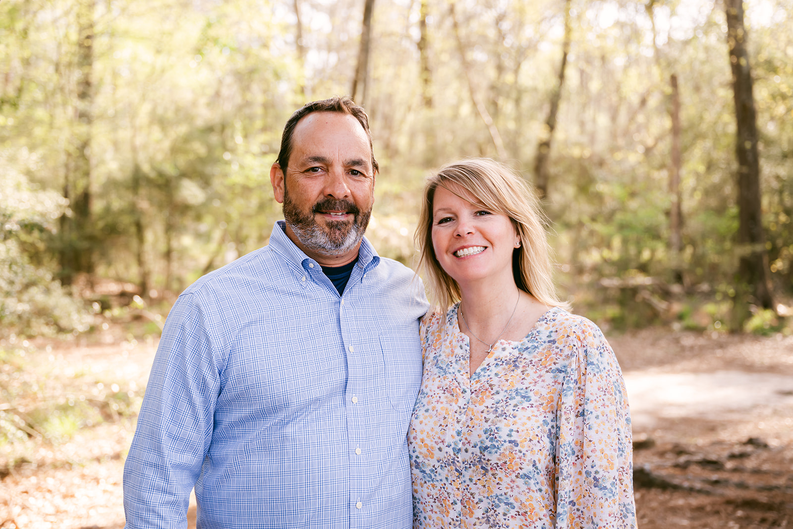 Parents of high school senior girl stand together in a wooded area with their arms around each other as they celebrate their daughter's accomplishment