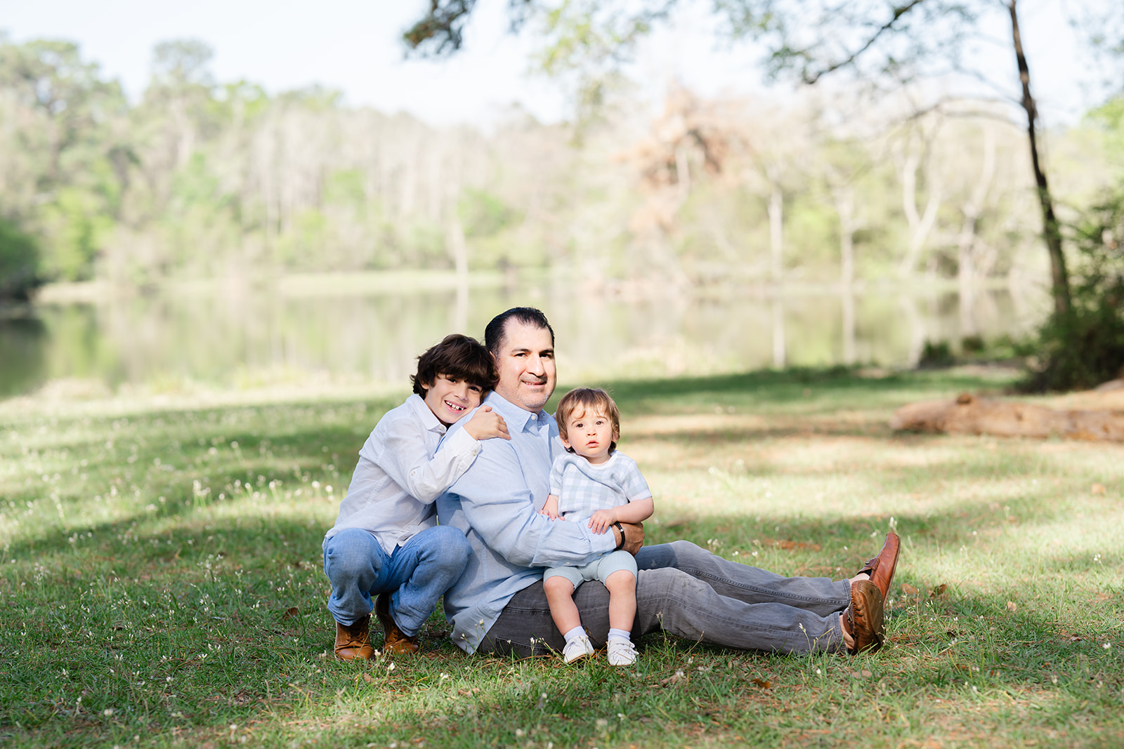 Dad seated on a grassy bank with his toddler son on his lap and his older son hugging him from behind