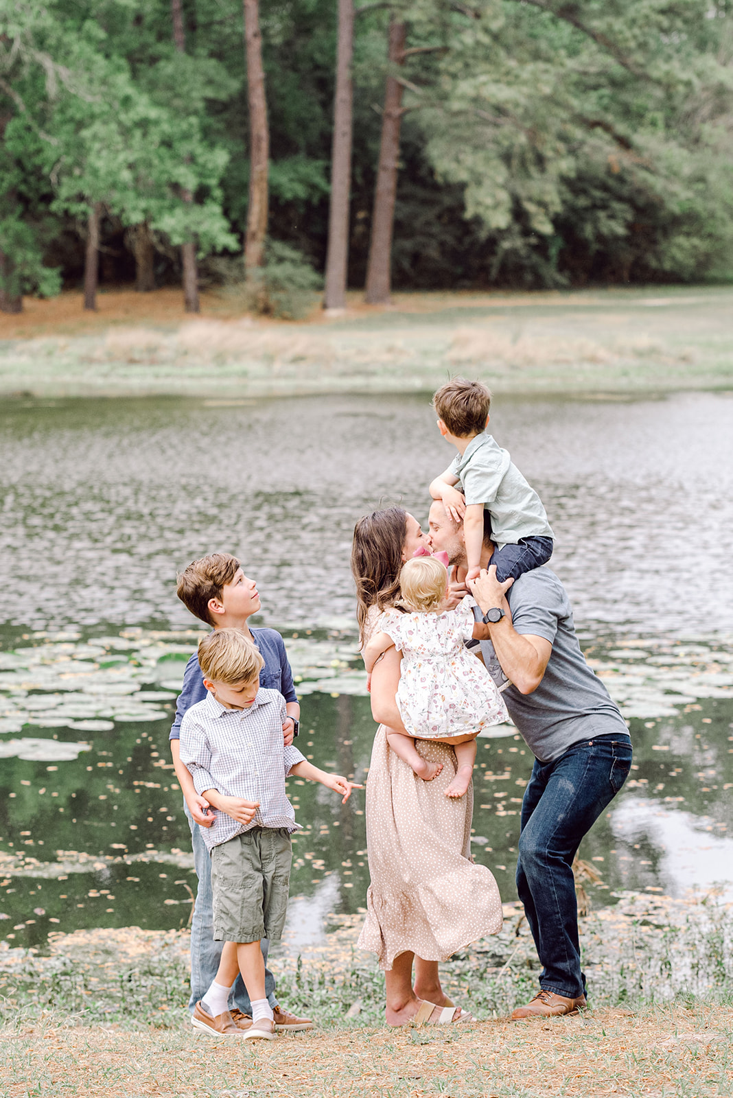 Dad with young boy on his shoulders and mom with baby in her arms exchange a kiss by the lake while their other children look on