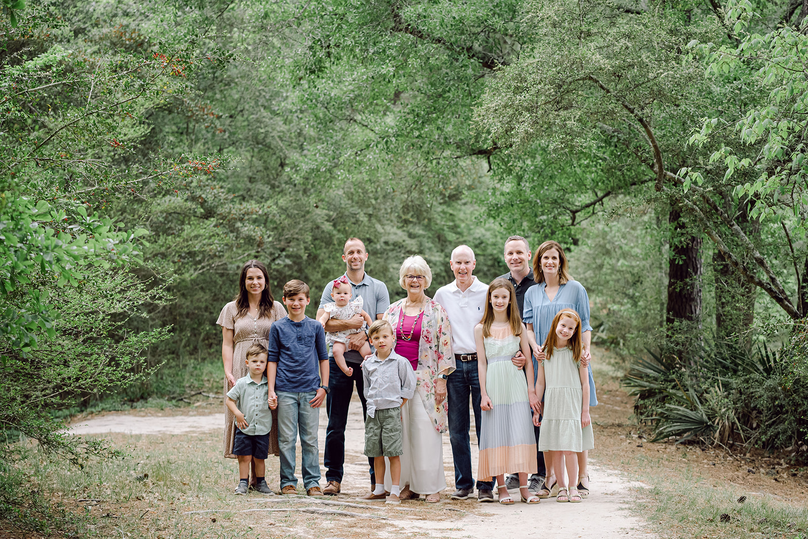 Grandparents with their children and grandchildren pose for an extended family photo in a wooded park