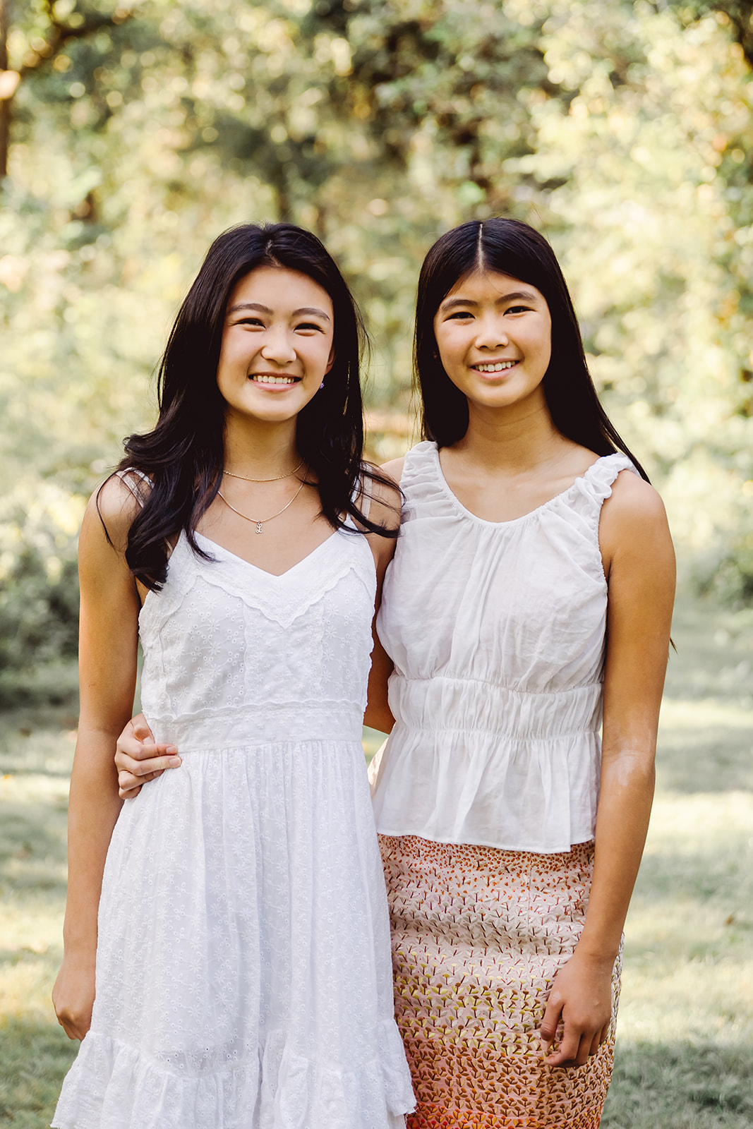 Teenage sisters in light-colored summer dresses stand with their arms around each other's waists during an outdoor Houston family photo session