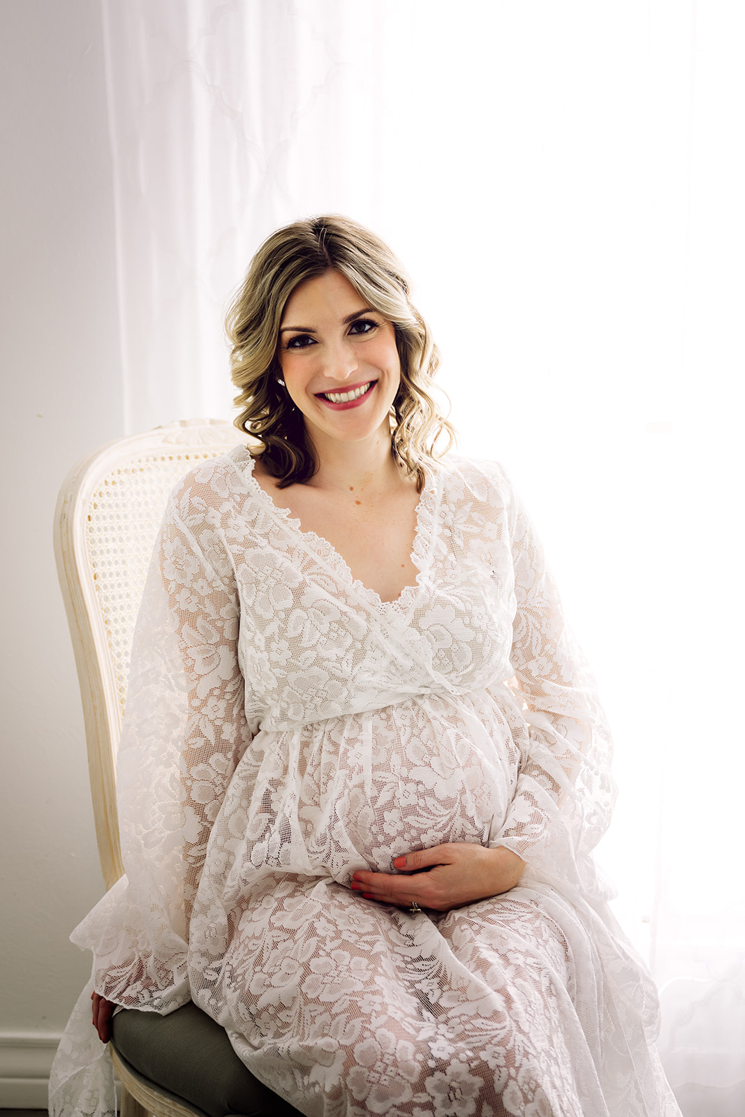 Woman in white lace maternity gown seated by a curtain window with natural light 