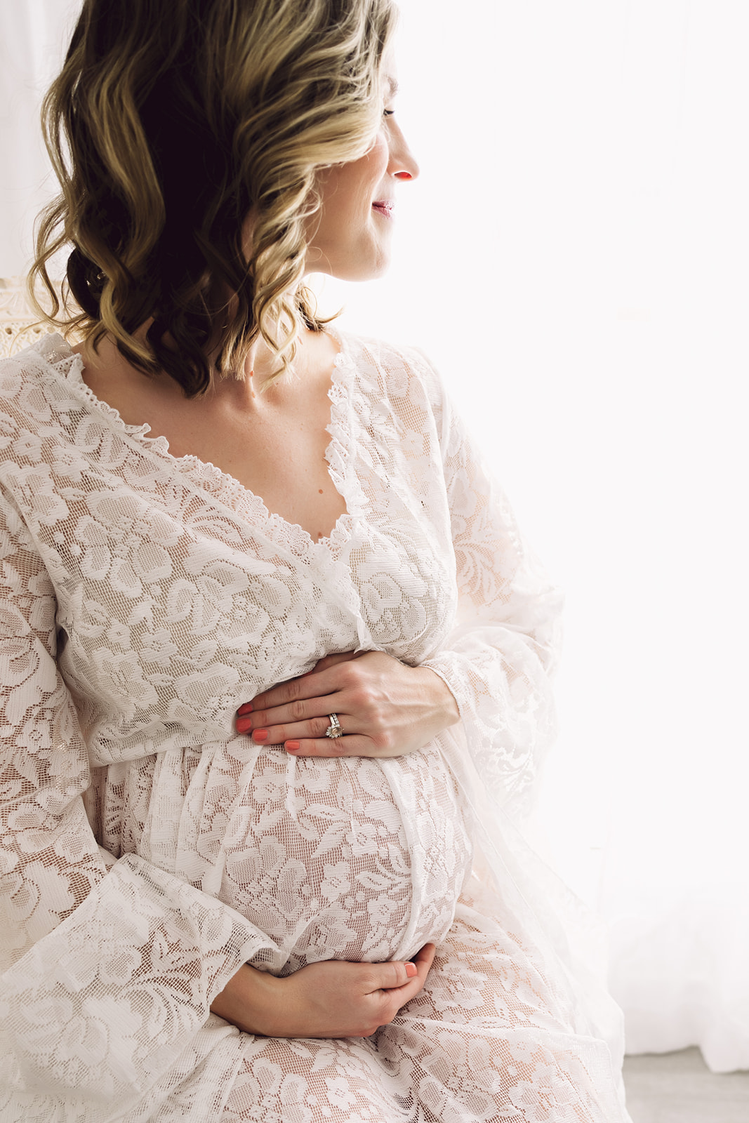 Woman in white lacy maternity dress gazes over her shoulder as her hands frame her baby bump