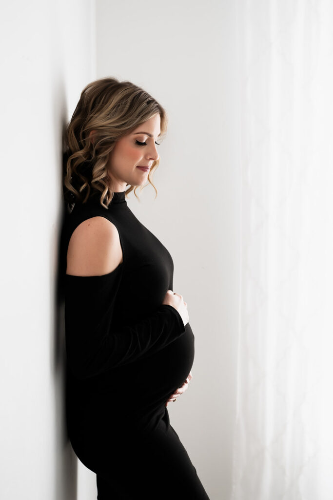 Woman in sleek black cold shoulder maternity dress, leaning back against a white wall during a studio maternity session in Houston Texas