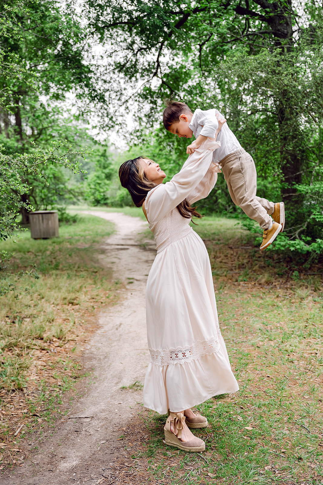 Pregnant mother in pink dress laughing as she lifts her toddler son into the air during an outdoor maternity session in Houston TX