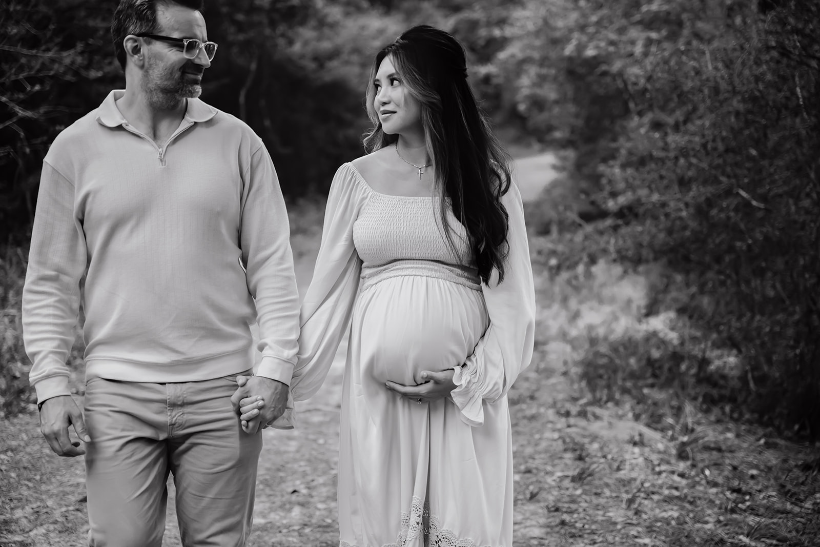 Pregnant woman in simple and flowing maternity gown smiles at her husband as they walk hand in hand down a wooded path together