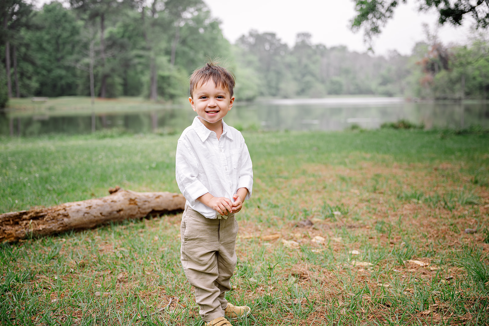 Smiling little boy in a button-up shirt and khaki pants standing by a pond during his mom's outdoor maternity session in Houston TX