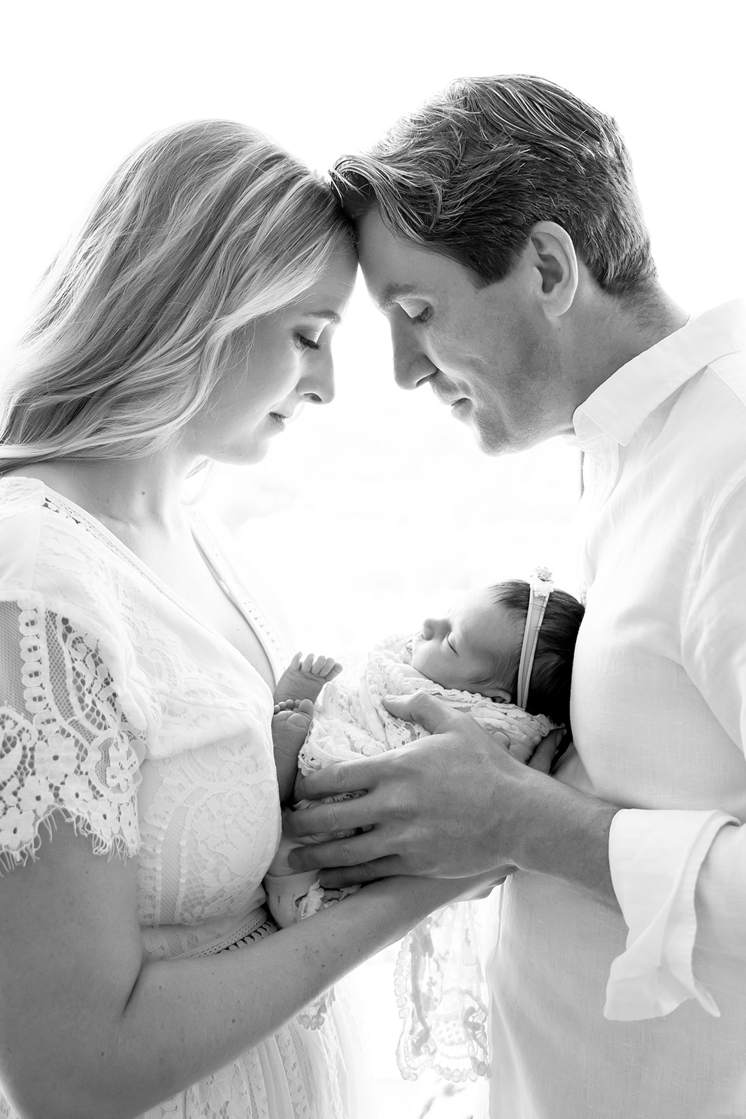 Backlit black and white image of mom and dad touching foreheads as they hold their newborn daughter between them