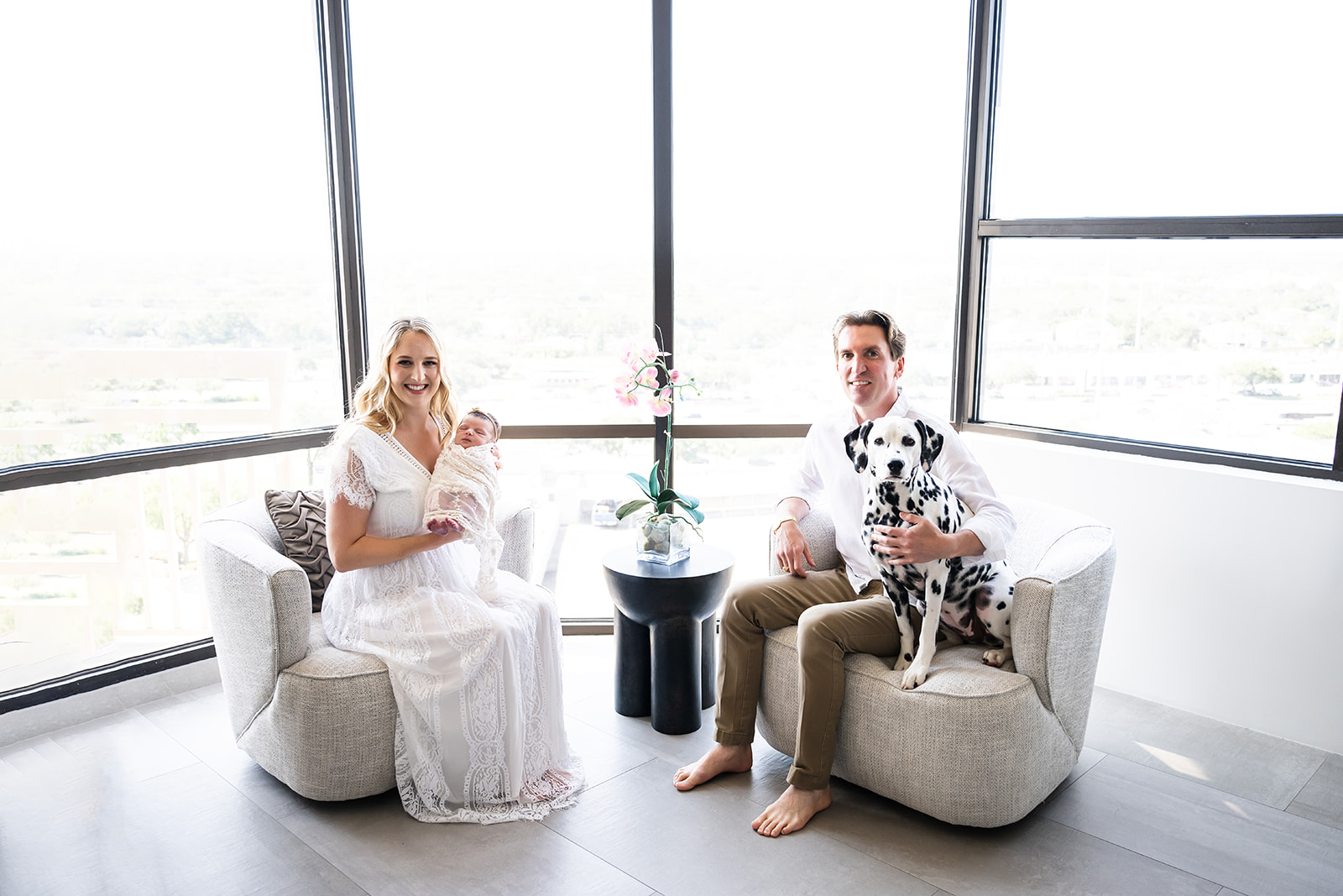 New parents seated by a window overlooking Houston Texas as mom holds their newborn and dad pats their Dalmatian pup.
