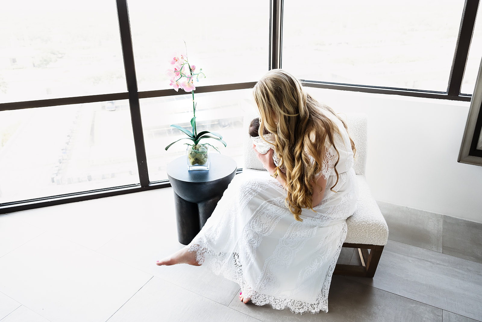 Young woman in white dress seated by a window leans forward to kiss her infant daughter during an at home newborn session in Houston Texas