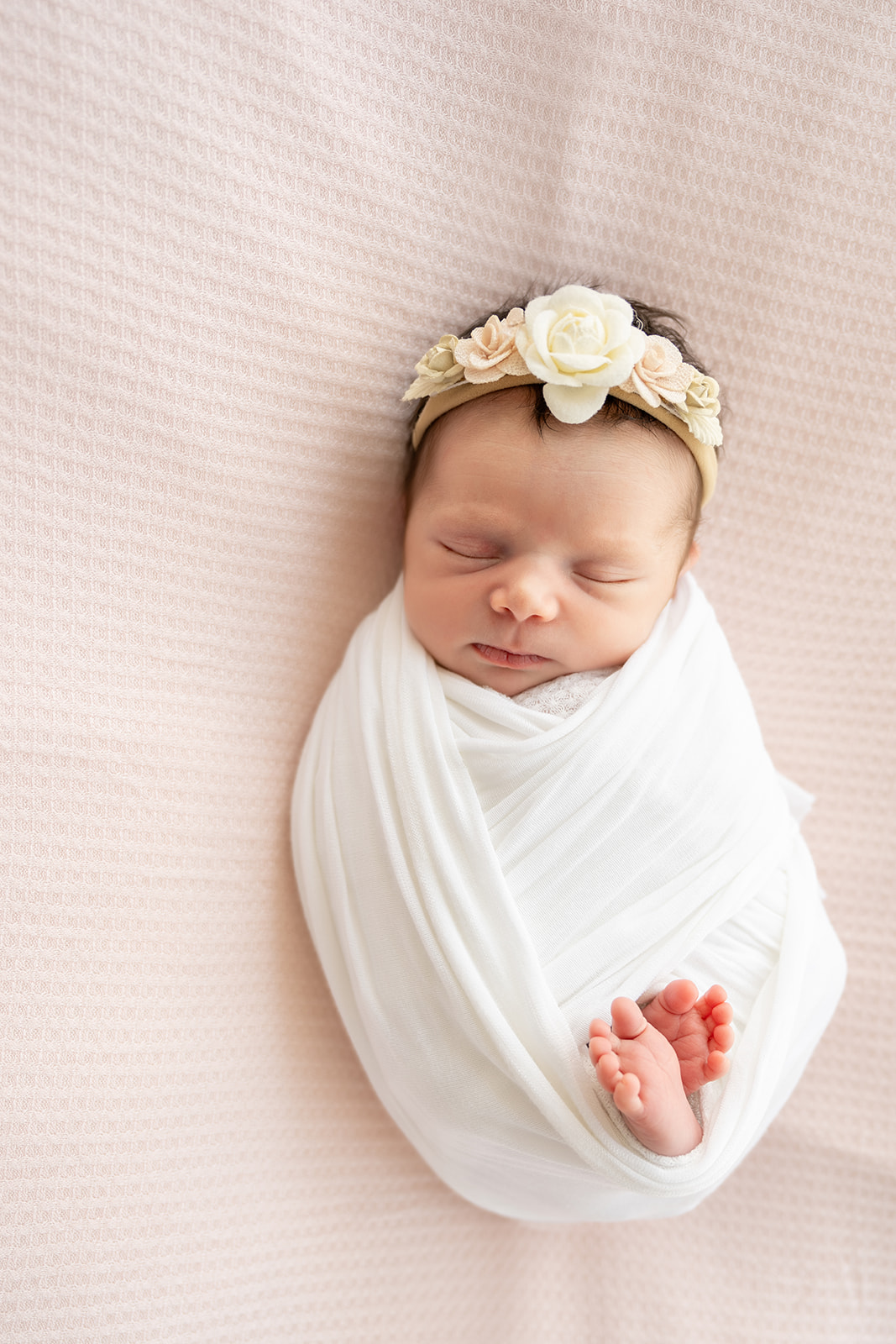 Newborn baby in white swaddle and floral headband sleeping peacefully on a pale pink background during her at home newborn session