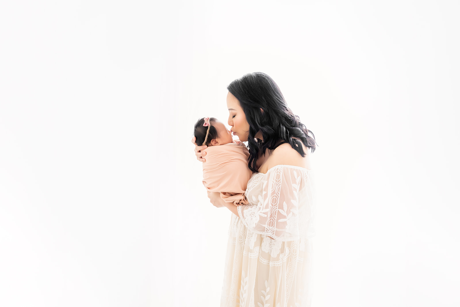 Mother in white dress leans forward to kiss her newborn baby girl as they stand against a natural light backdrop in a Houston studio