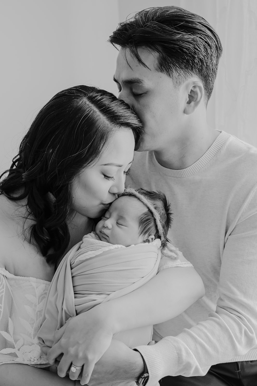 Black and white family portrait of mother leaning down to kiss her newborn while her husband wraps his arms around them both