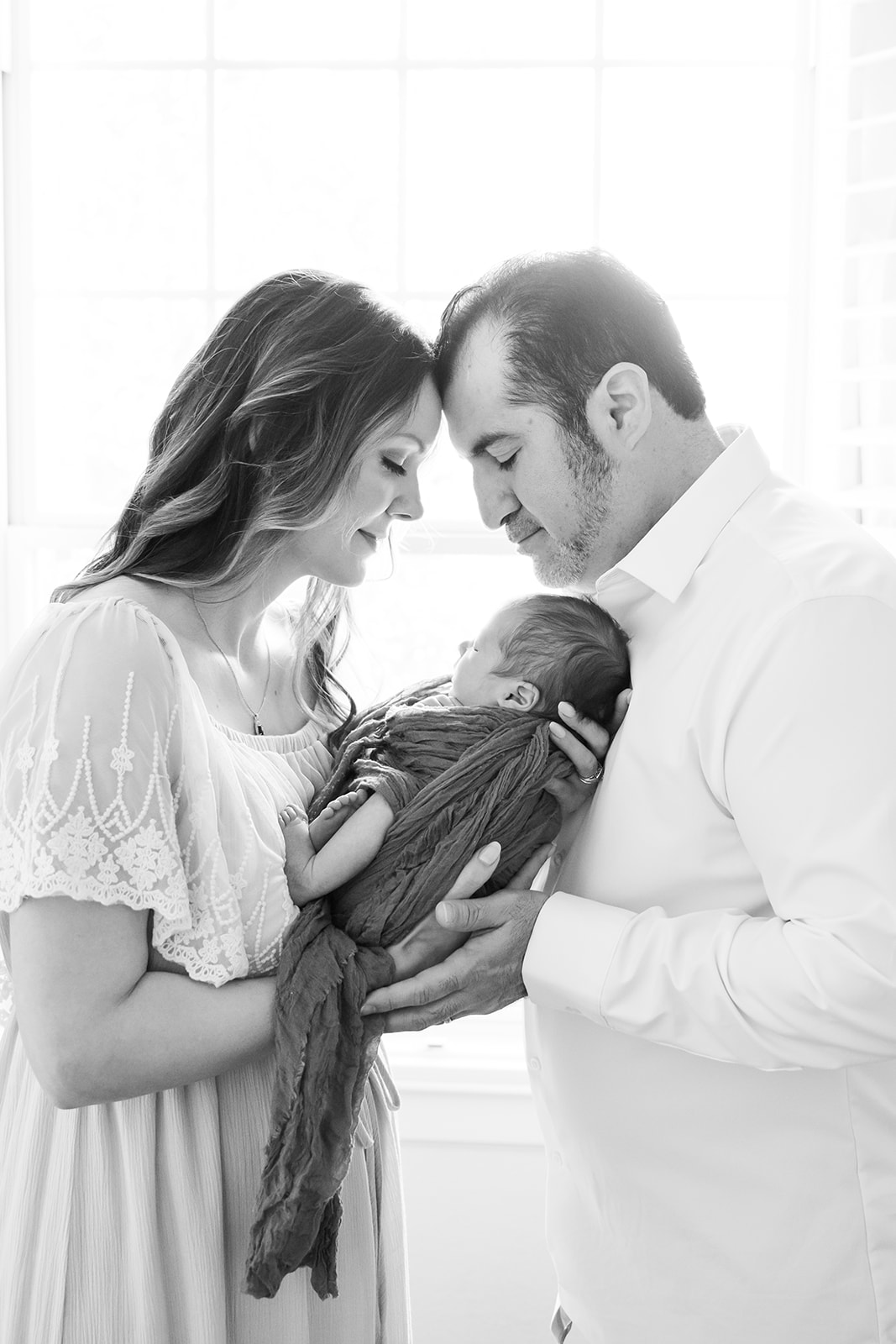 Black and white image of parents holding their newborn between them and touching foreheads as they stand in front of a window