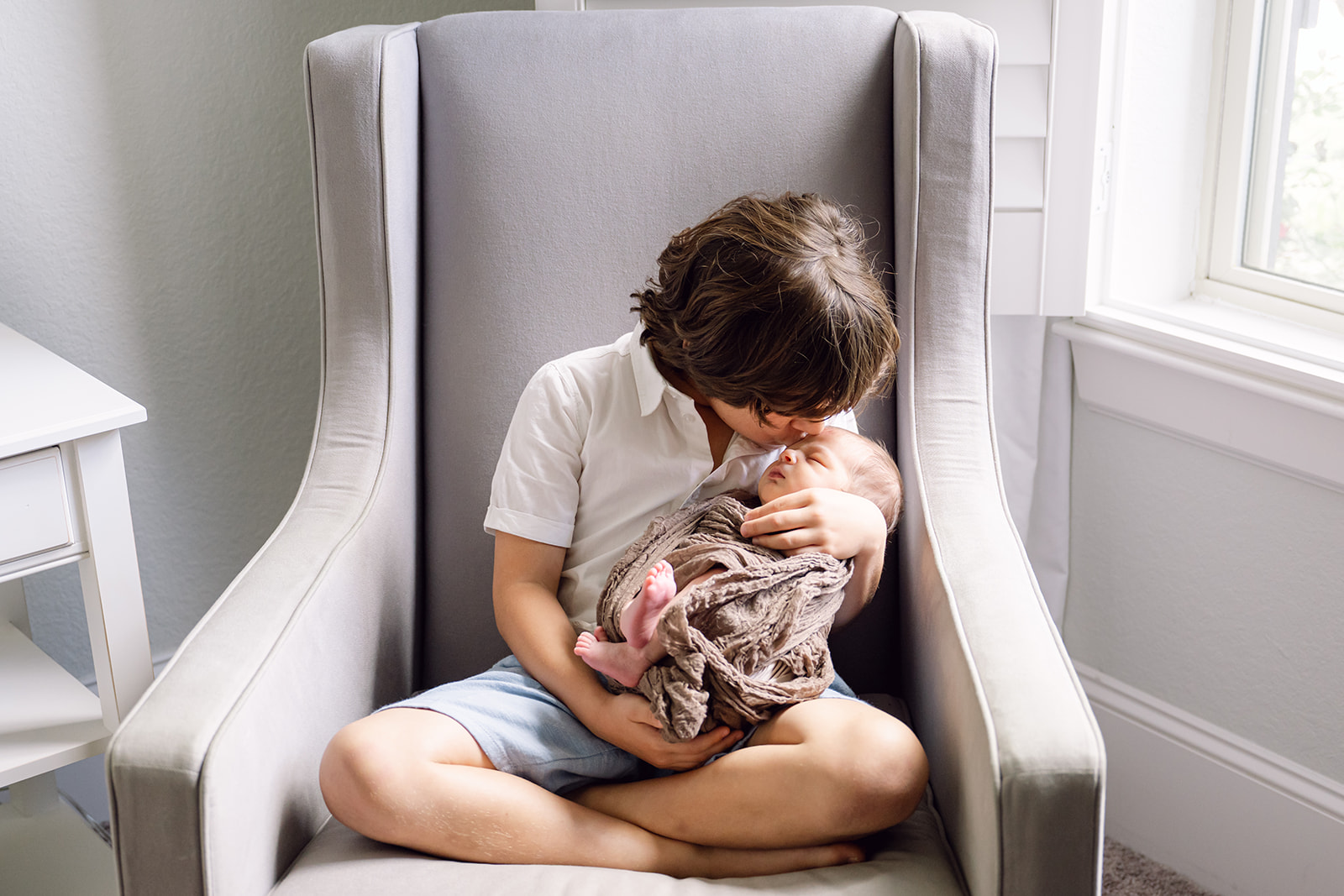 Little boy in gray chair by a window holding his infant brother and giving him a kiss on the forehead