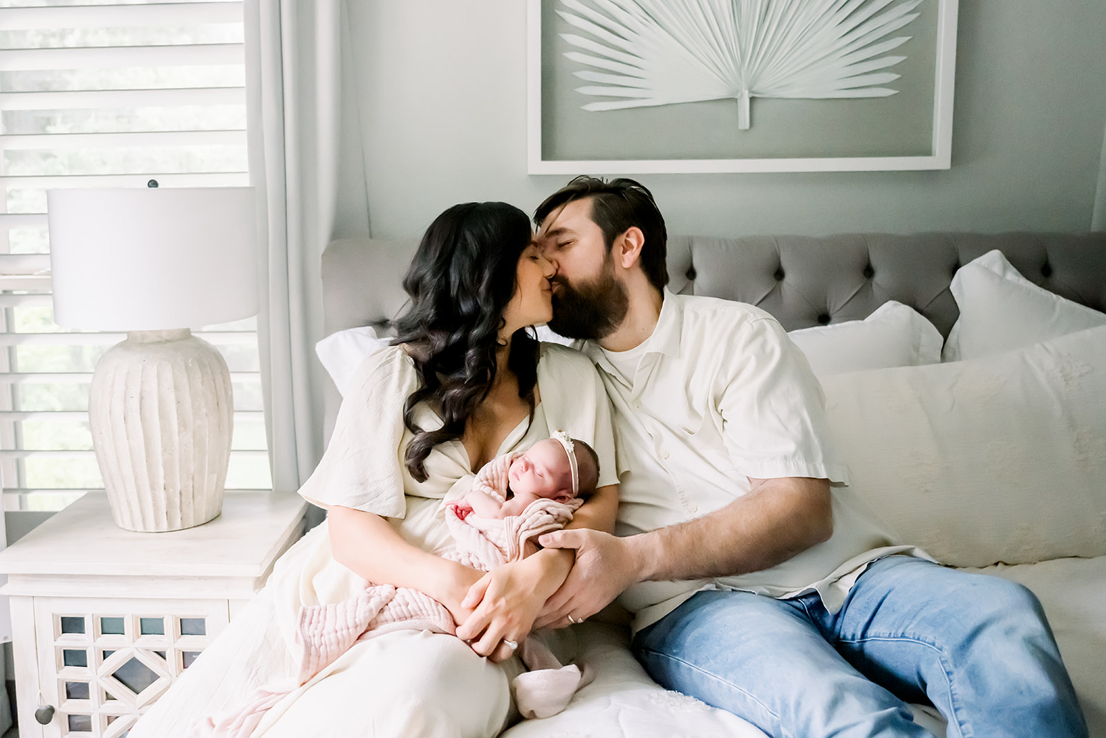 Mom and Dad exchange a kiss as they sit on the bed together holding their newborn between them