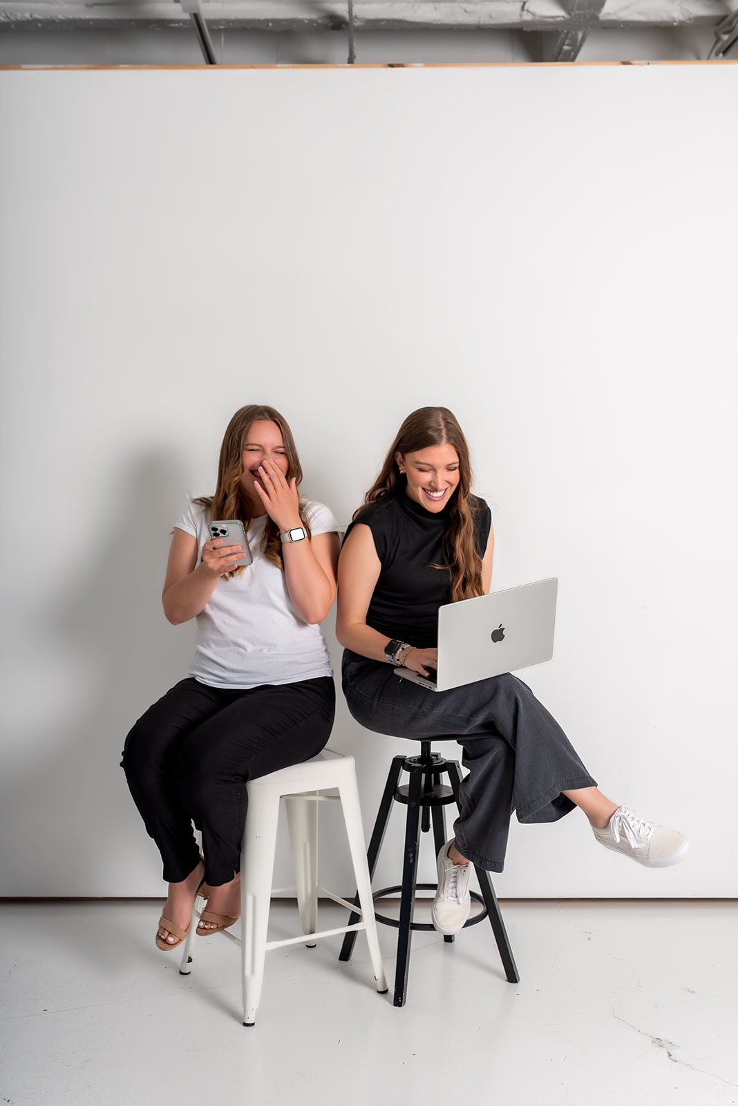 Two female coworkers laughing together as they work, one on her phone and the other on a laptop