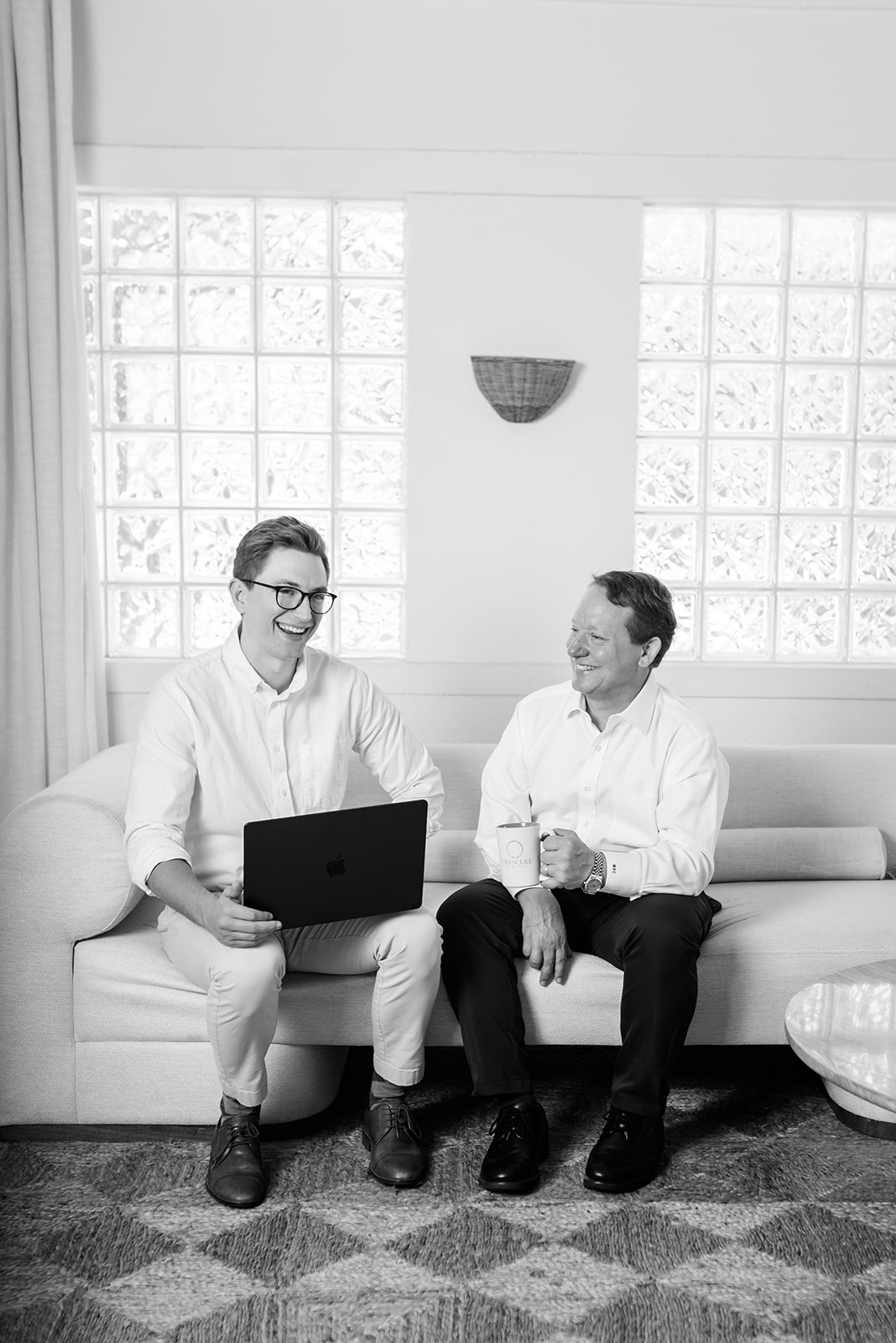 Two men laughing as they sit together on a couch, one with a laptop and one with a mug of coffee in hand, during a business meeting break