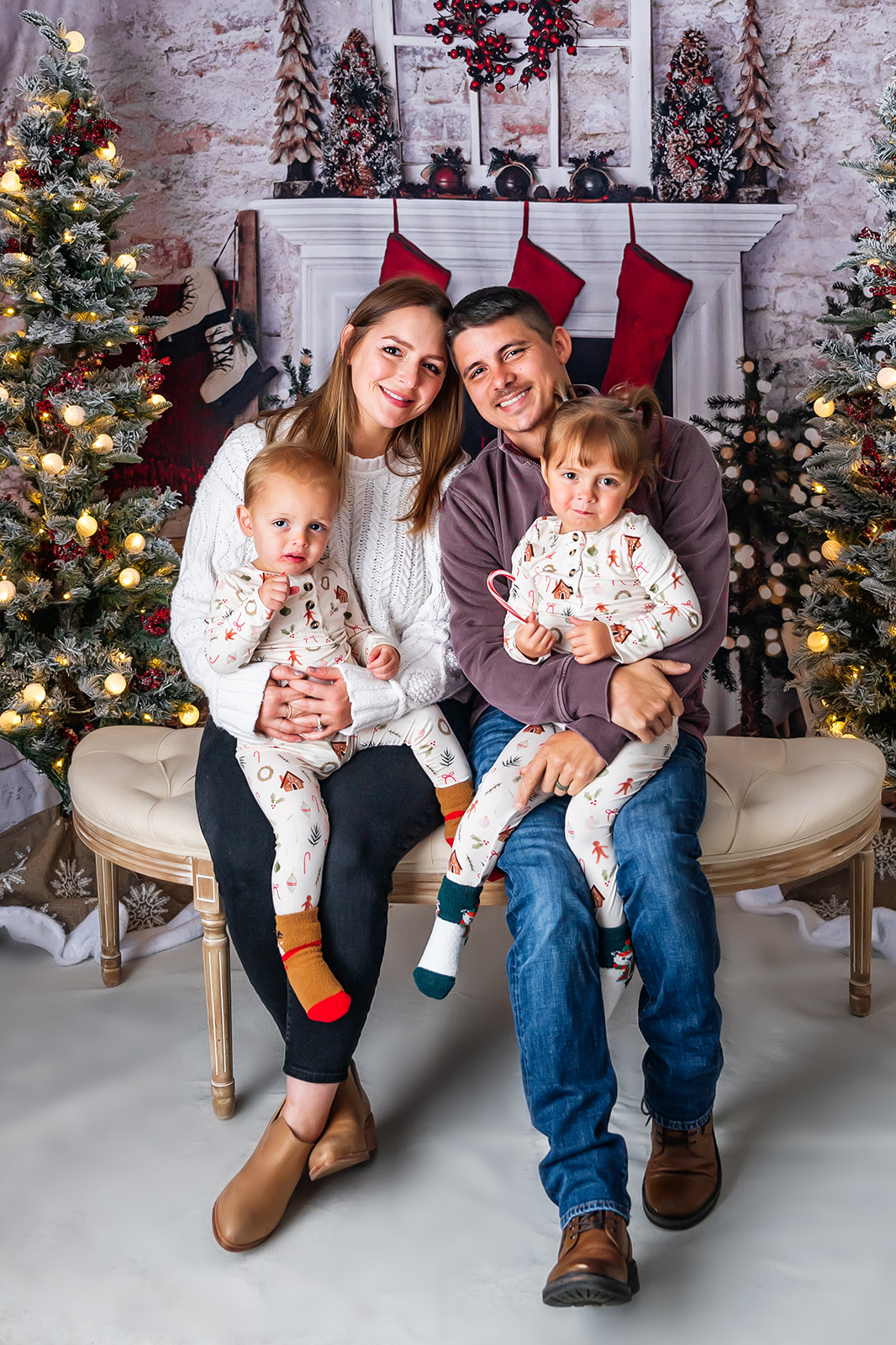Family seated on white bench in front of a fireplace hung with stockings and a decorated Christmas tree in the background