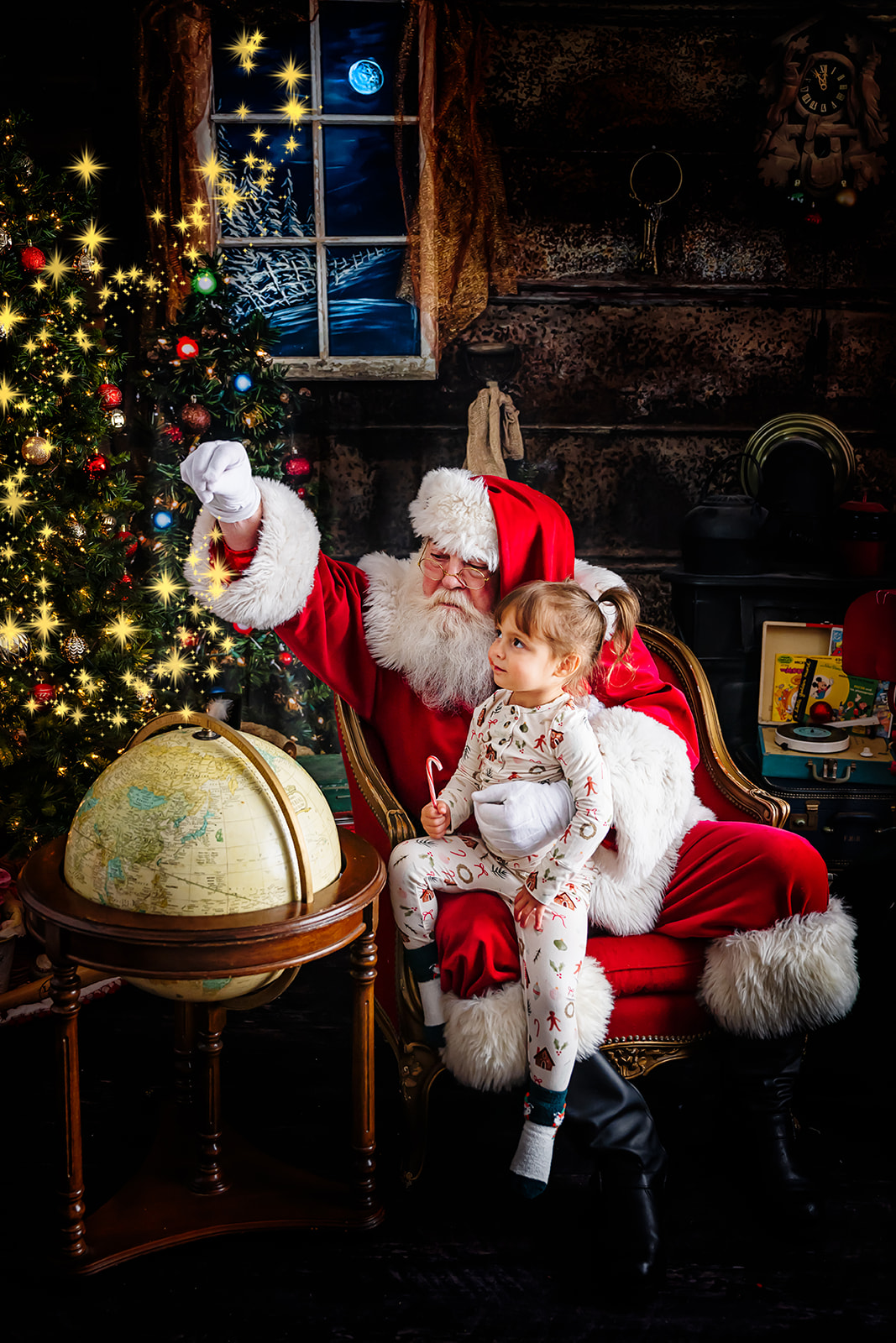 Santa holding a toddler girl on his knee while they look at a floor globe together
