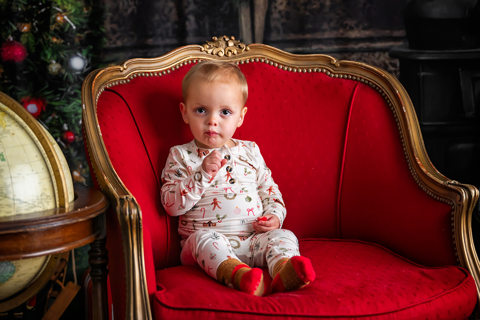 Toddler boy in holiday pajamas seated on a red upholstered wingback chair with a Christmas tree in the background