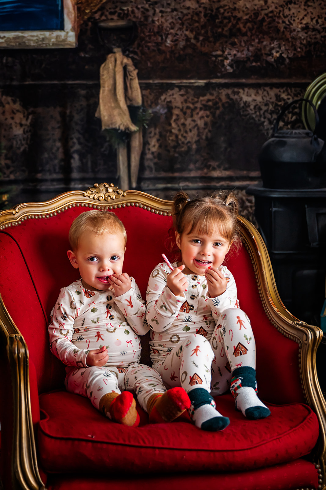 Siblings in festive PJs seated on a red and gold upholstered chair while they enjoy peppermint candy canes
