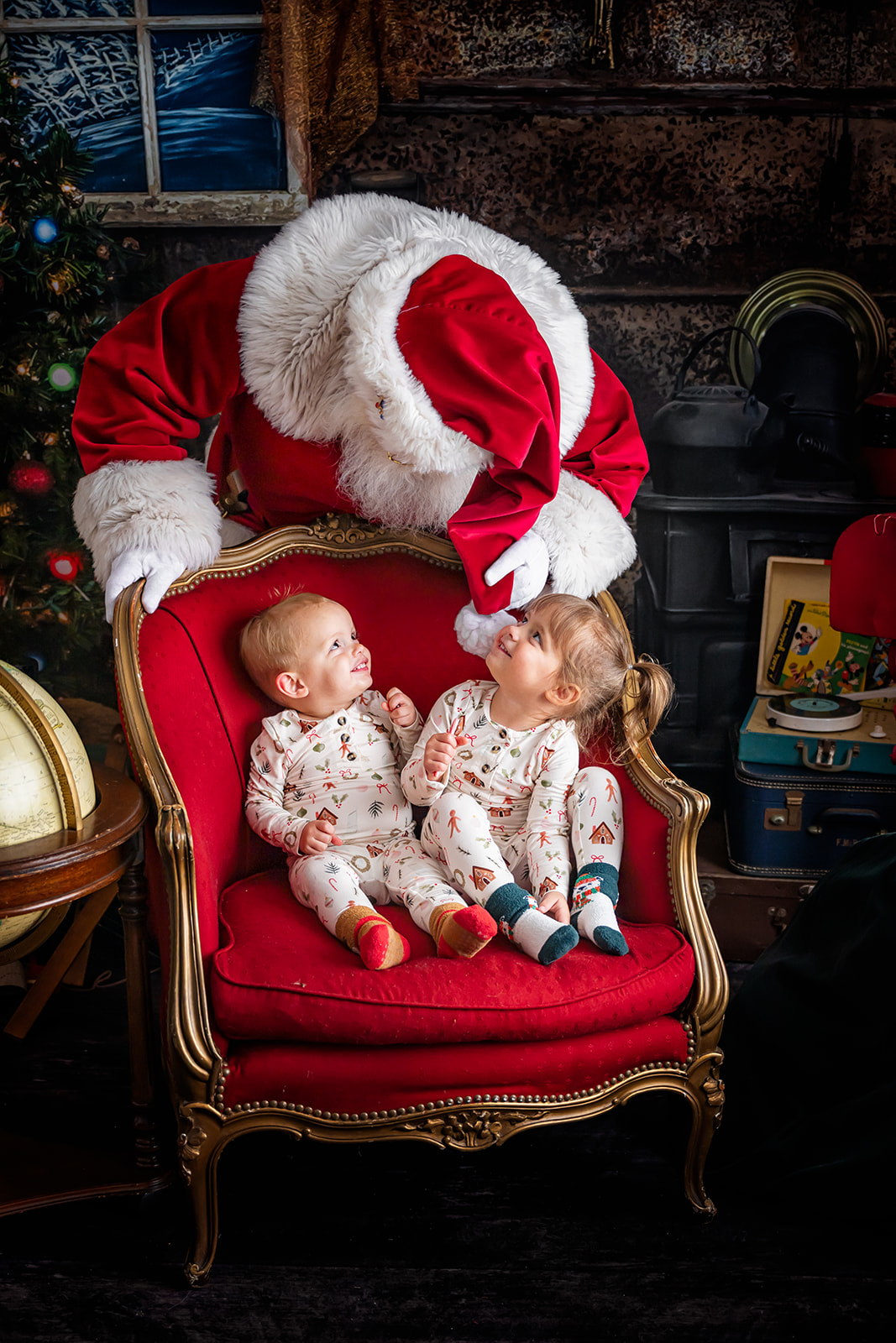 Toddler siblings smiling up at Santa as he leans over their chair 